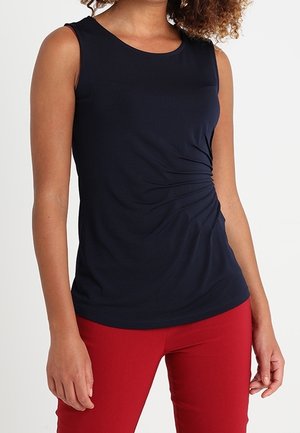 Vrouw draagt een mouwloze marineblauwe top met zijsamenspel en een rode broek, staand tegen een effen witte achtergrond.