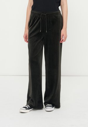 Personne portant un pantalon ample en velours noir avec une taille à cordon et des baskets en toile noires et blanches, debout sur un sol blanc.
