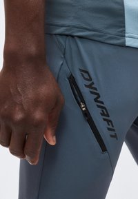 Κοντινό σε χέρι ατόμου δίπλα σε γαλάζιο-γκρι σορτς με μαύρη τσέπη με φερμουάρ και λογότυπο "DYNAFIT" στο μπούτι.