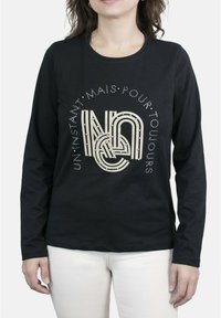 Camisa negra de manga larga con un diseño gráfico texturizado en crema y tachuelas decorativas. Se lleva con pantalones de color claro.