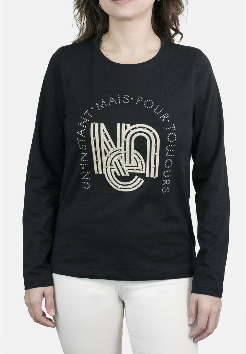 Camisa negra de manga larga con un diseño gráfico texturizado en crema y tachuelas decorativas. Se lleva con pantalones de color claro.
