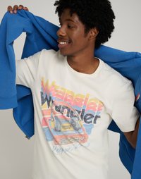 Λευκό T-shirt με πολύχρωμο σχέδιο ενός ρετρό κασετόφωνου με ακουστικά. Ένα μπλε ριχτό μπουφάν είναι περασμένο στον έναν ώμο.