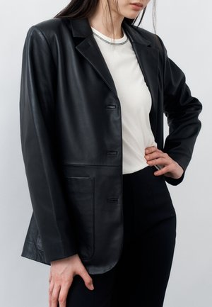 Femme portant un blazer en cuir noir sur un t-shirt blanc et un pantalon noir, posant avec une main sur la hanche et l'autre détendue.