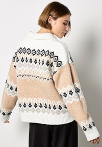 Kremowy i beżowy sweter dziany z geometrycznymi czarno-białymi wzorami, luźny krój, ściągacze na mankietach i kołnierz złożony na pół. Teksturowane wykończenie.