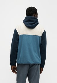 Vīrietis ar īsiem cirtainiem matiem, valkājot krāsu blokā iedalītu flīsa jaku ar tumši zilu kapuci, krēmkrāsas augšējo daļu un zilu apakšējo muguras daļu, skatoties prom.