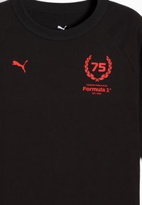 Mustapaitainen puuvillapaita, jossa on punainen graafinen teksti "75" laakerinoksalla, "PASSION THEN & NOW" ja "Formula 1" -logo vasemmalla puolella. Puma-logo punaisena hihassa.