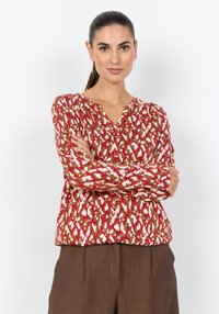Rote und cremefarbene Bluse mit Muster, V-Ausschnitt und langen Ärmeln, aus leichtem Stoff; kombiniert mit braunen Hosen.