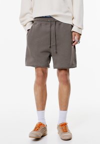 Graue Baumwollshorts mit elastischem Taillenbund und Kordelzug, mit ausgefransten Säumen. Getragen mit beigen Sneakers und weißen Socken.