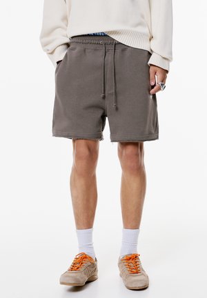 Pantaloni corti in cotone grigio con una vita elasticizzata e lacci, caratterizzati da orli sfrangiati. Indossati con sneakers beige e calze bianche.
