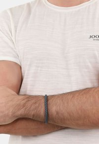Pulsera trenzada de cuero negro en una muñeca, contrastando con una camiseta de textura clara y de manga corta. Diseño simple y minimalista.