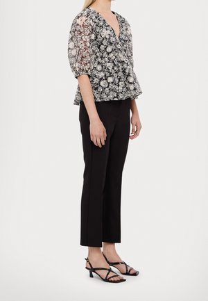 Femme portant un pantalon court noir, des sandales à talons bas noires à brides, et un chemisier transparent noir et blanc à motifs floraux avec des manches bouffantes.