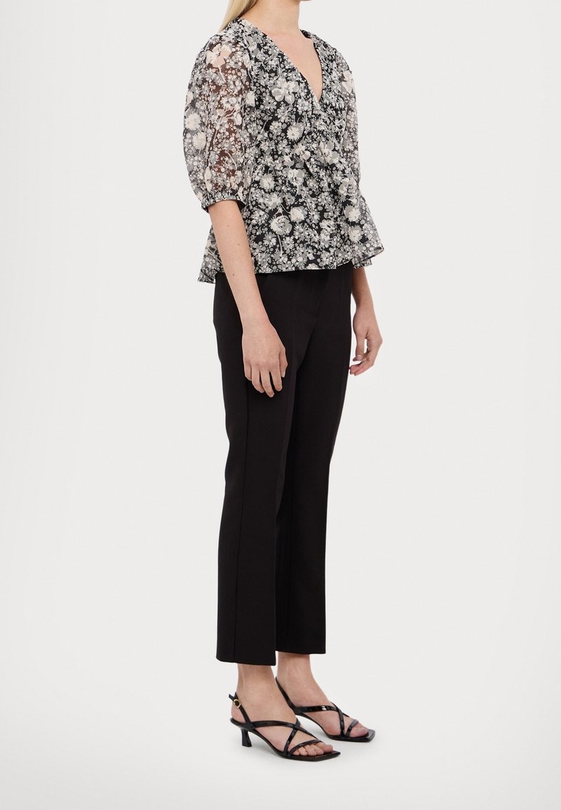 Femme portant un pantalon court noir, des sandales à talons bas noires à brides, et un chemisier transparent noir et blanc à motifs floraux avec des manches bouffantes.