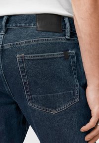 Jeans de mezclilla azul oscuro con un parche de cuero en la cintura, que presentan costuras blancas y una pequeña etiqueta negra en el bolsillo trasero.