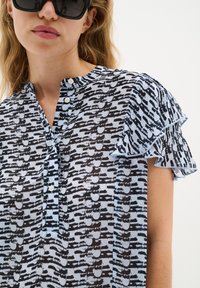 Blusa a maniche corte in tessuto fantasia, con un design nero e azzurro chiaro, colletto con bottoni e bordi delle maniche arricciati.