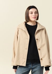 Beżowa wodoodporna kurtka z kołnierzem, zatrzaskami i luźnym krojem. Noszona na czarnym swetrze, w zestawieniu z jasnoniebieskimi dżinsami.