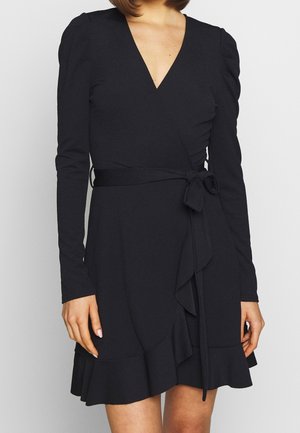 Nainen pukeutuneena pitkähihaiseen mustaan kietaisumekkoon, jossa on V-aukko, röyhelöhelma ja vyötäröllä solmittu rusetti, yksinkertaista vaaleaa taustaa vasten.