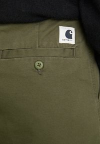 Zaļās auduma bikses ir aprīkotas ar priekšējo kabatu, kurai ir atbilstoša pogas, mazu baltu etiķeti ar logotipu un gludu tekstūru.