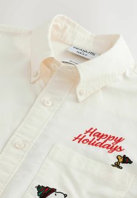 Beige Hemd mit Knöpfen, auf dem "Frohe Feiertage" in Rot gestickt ist, mit Snoopy und Woodstock, die grüne und rote gestreifte Hüte tragen.