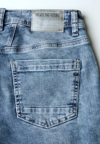 Světle modré džíny z denimu, které mají zadní kapsu s kontrastním stehem a obdélníkovou koženou nášivku připevněnou na pase.