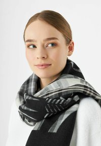 Jonge vrouw met lichte huid en bruin haar draagt een zwart en grijs geruite sjaal over een witte top en kijkt iets opzij.