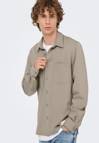 Beige overhemd met lange mouwen, knoopsluiting, een borstzak en een getailleerde kraag. Gedragen over een wit T-shirt, getoond op een mannelijk model.