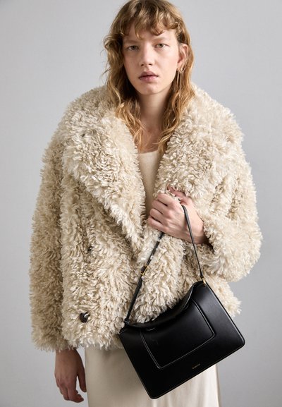 Beige nepgaren jas met een gestructureerde afwerking, gecombineerd met een zwarte leren handtas met witte stiksels en een afneembare schouderband.