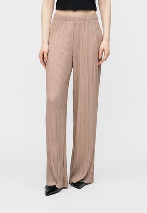 Kvinde iført beige plisserede bukser med høj talje og vide ben, kombineret med sorte spidse sko og en sort cropped top.