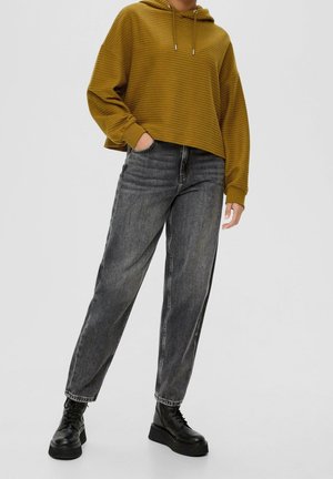 Person iført okkergul ribstrikket hættetrøje, falmede sorte jeans med høj talje og sorte platform-sko med snørebånd, stående med hånden i lommen.