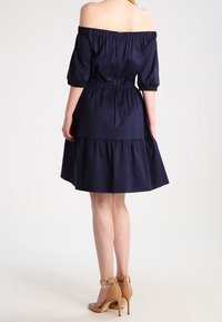 Marineblaues Off-Shoulder-Kleid mit Puffärmeln und elastischem Taillenbund. Gestufter Rockdesign. Getragen mit braunen High Heels.