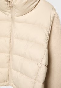 Beżowa, krótką kurtka z ocieplaną, gładką powłoką oraz ściągaczowymi rękawami. Posiada pełny zamek błyskawiczny oraz kaptur.