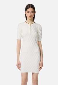 Weißes Strickkleid mit strukturiertem Rautenmuster, kurzen Ärmeln, zwei Vorderentaschen und einem mit Gold akzentuierten Kragen mit einem zentralen Reißverschluss.