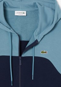 Bluza z kapturem z mieszanki jasnoniebieskiego i granatowego materiału, z zapięciem na zamek błyskawiczny oraz małym haftowanym logo krokodyla na piersi.