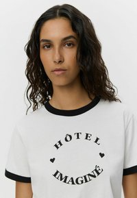 Vit T-shirt med svart ringkrage, med texten 'HÔTEL IMAGINÉ' och svarta hjärt-accenter. Modellen har vågigt hår som hänger ner.