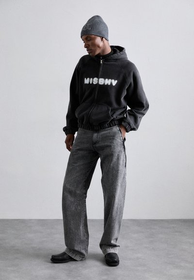 Schwarze Zip-Up-Hoodie mit "MISBHV" in weiß bedruckt, kombiniert mit lockeren grauen Jeans und einer grau gestreiften Mütze. Schwarze Schuhe runden das Outfit ab.