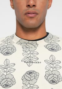 Suéter de color claro con un gran patrón floral en gris, con un cuello redondo y el logotipo "marimekko" impreso en el centro debajo del diseño.