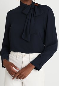 Nainen, jolla on yllään tummansininen pitkähihaisen pusero, jossa on solmittava kaulus, sekä valkoiset korkeavyötäröiset housut, kädet rennoisti ristissä edessä.