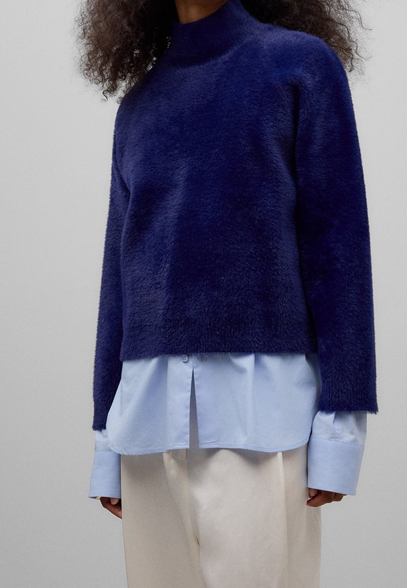 Marineblå sweater med pelsagtig tekstur, høj hals og lange ærmer, lagdelt over en lyseblå skjorte med knapper. Blød tekstur og kasseformet silhuet.