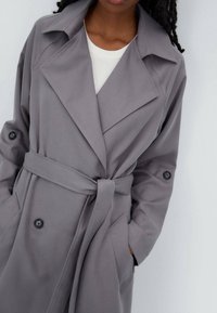 Grauer Trenchcoat mit tailliertem Gürtel, großem Kragen und Knopfdetails. Aus glattem Stoff gefertigt, mit Fronttaschen und lässiger Passform.