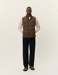 Brūns pūkains bezmugurkaula veste ar augstu apkakli un priekšējo rāvējslēdzēju. Uzvilkta pār gaišas krāsas biezas adītas džemperis un kombinēta ar tumšiem biksēm.