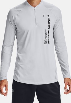 Mann trägt ein hellgraues, langärmeliges Under Armour Shirt mit Viertelreißverschluss und schwarzem Textlogo auf der Vorderseite, kombiniert mit schwarzen Shorts.