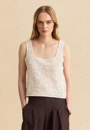 Mujer con cabello largo y rubio que lleva una blusa sin mangas de encaje beige y pantalones de talle alto de color marrón oscuro, manos en los bolsillos, fondo neutro.