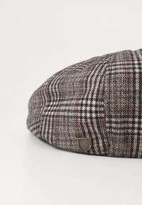 Flat cap made of plaid fabric in gray, black, and red. Features a small metal emblem on the side. Round, structured design with texture.