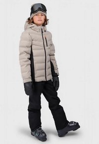 Barn som har på seg en beige boblejakke, svarte snøbukser, svarte hansker, grå skistøvler, lue og skibriller på hodet, står foran en ensfarget bakgrunn.