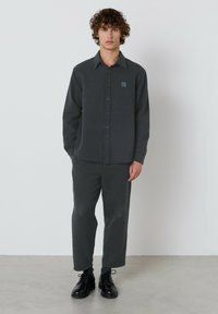 Mladenič stoji ob beli steni, oblečen v temno sivo srajco na gumb, usklajene hlače, črne vezalke in sive nogavice.