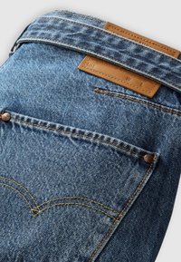 Vaqueros de mezclilla azul con una etiqueta de cuero, costuras en contraste y un detalle de botón de metal. Presenta un bolsillo trasero y una forma clásica de bolsillo.