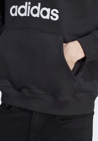 Crna majica s kapuljačom s bijelim "adidas" logom na prsima, s prednjim džepom i rebrastim manžetama i porubom. Mekani materijal od mješavine pamuka.