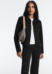 Femme portant une veste zippée noire, un haut blanc, un pantalon noir, et portant un sac à bandoulière beige à carreaux sur un fond blanc uni.