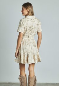Abito beige con maniche corte, ricamo floreale, orlo svasato, vita cintrata e colletto. Abbinato a stivali alla caviglia con frange color sabbia.