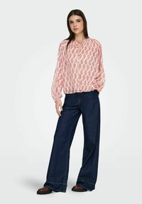 Blouse rose clair à motif de feuilles, avec un col noué et une fermeture boutonnée sur le devant, associée à un jean large bleu foncé et des bottes marron.