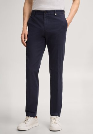 Mężczyzna noszący granatowe dopasowane spodnie, białe trampki i białą koszulę, stojący z jedną ręką w kieszeni.